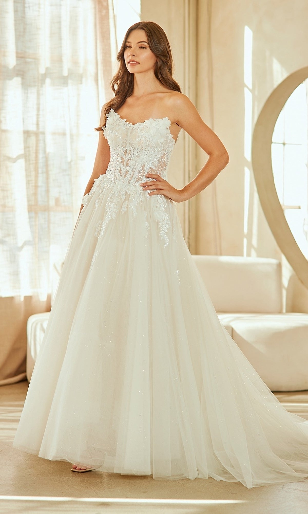 A woman stands indoors wearing the Amelia Couture 2141, a strapless long wedding dress with delicate lace details and a voluminous glitter tulle skirt for a dreamy, floor-length bridal look.